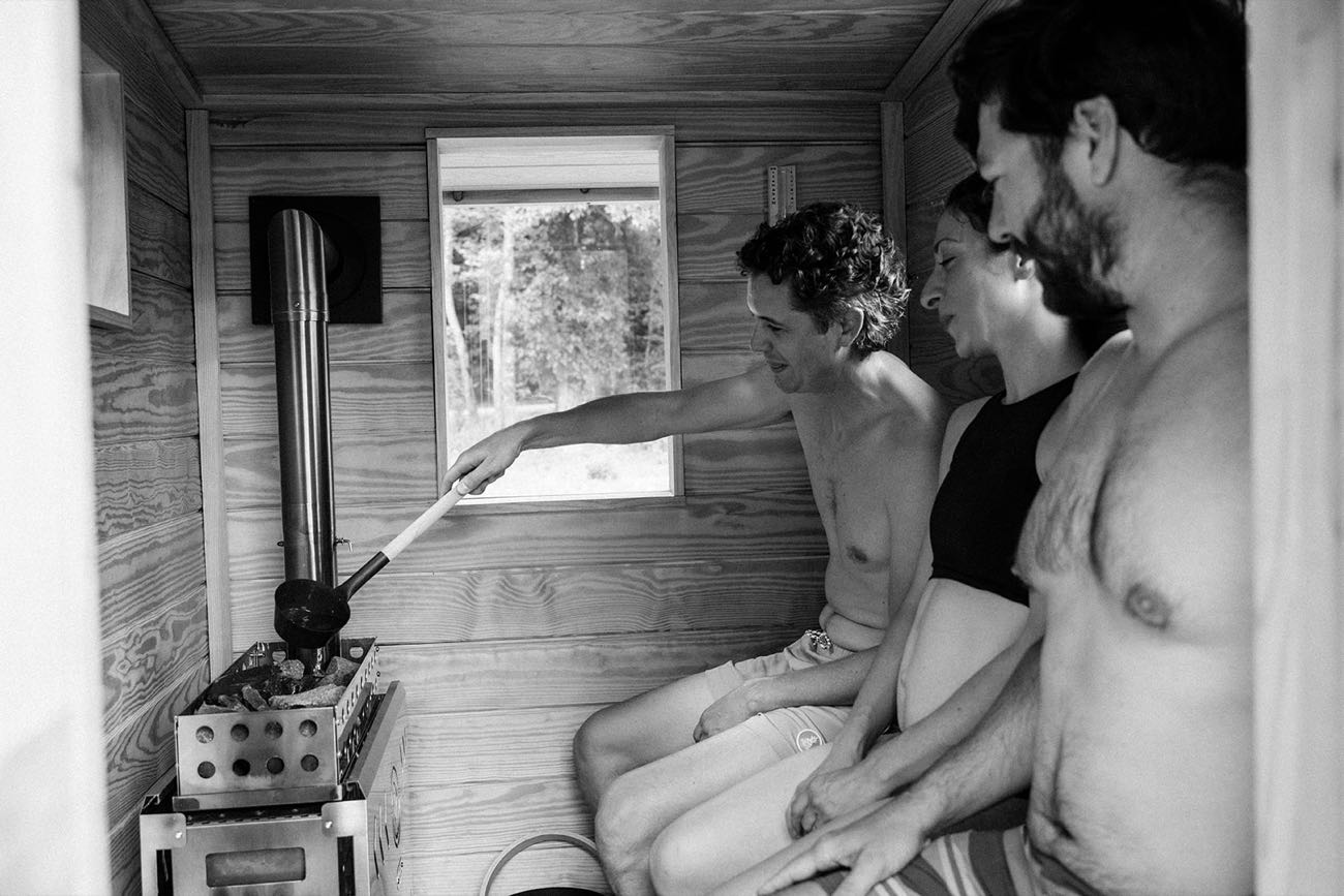 A ladle of water poured over the hot stones as three people sit along the sauna bench