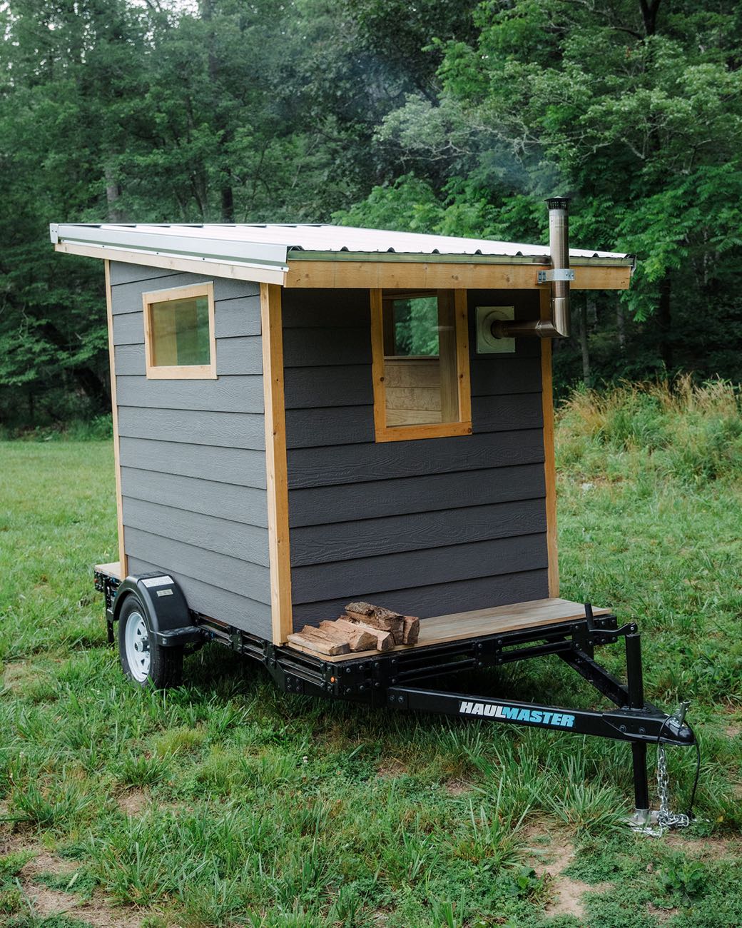 Three-quarter view of the Bushlot sauna on its trailer, light wood corner trim and window framing visible, split firewood stacked on the deck