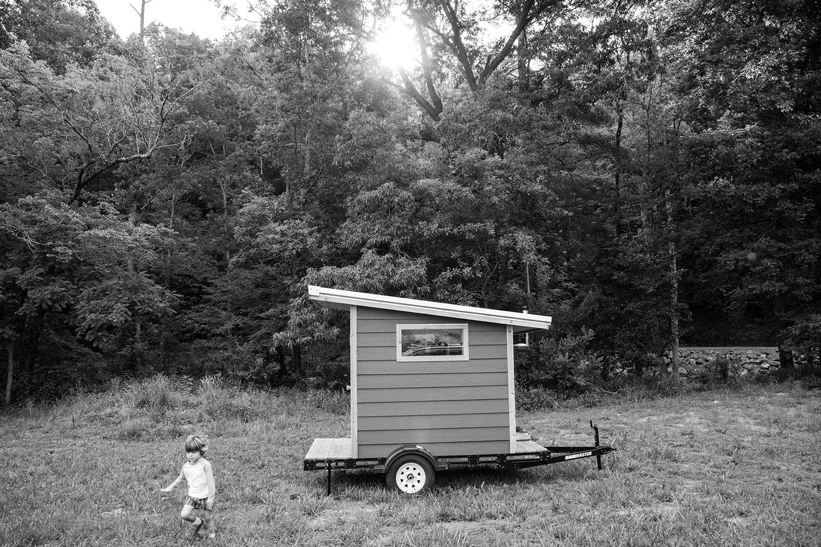 Wide view of the sauna alone in a mown field, sun flaring through the treeline, a small child walking across the grass