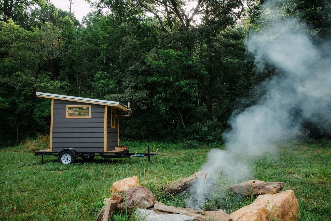 The sauna in a field at dusk with a fire pit burning in the foreground and smoke drifting across the trees