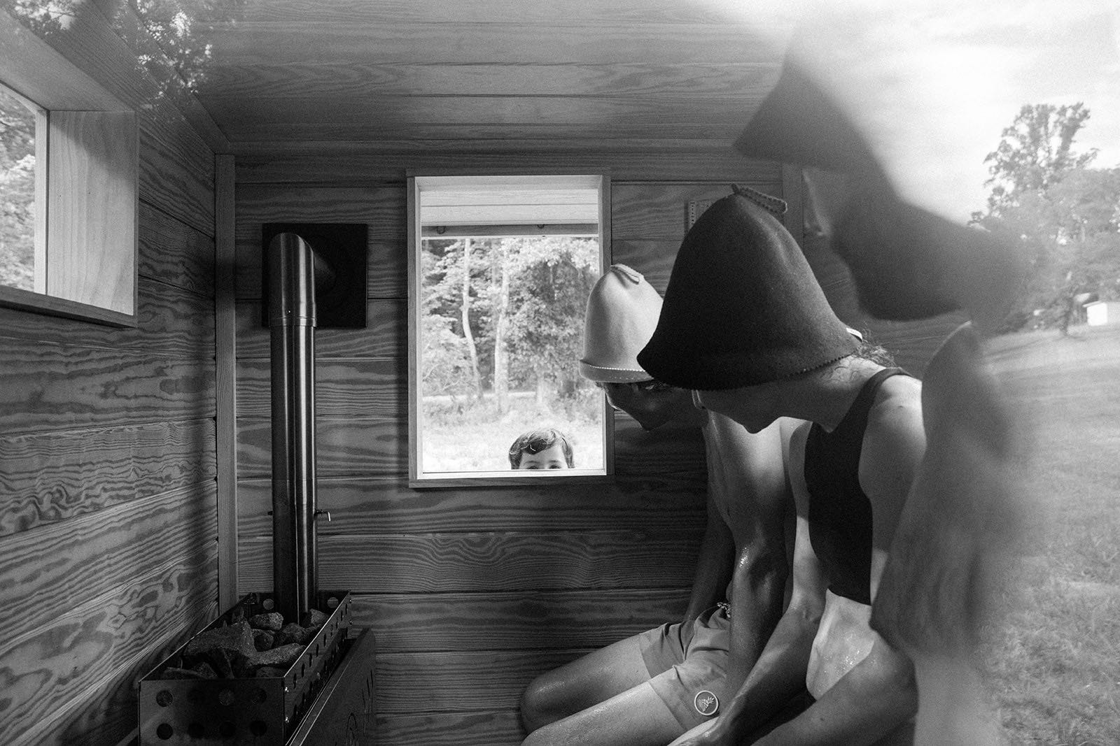 Inside the sauna, two people in wool sauna hats on the bench while a child peers in through the window from the field outside