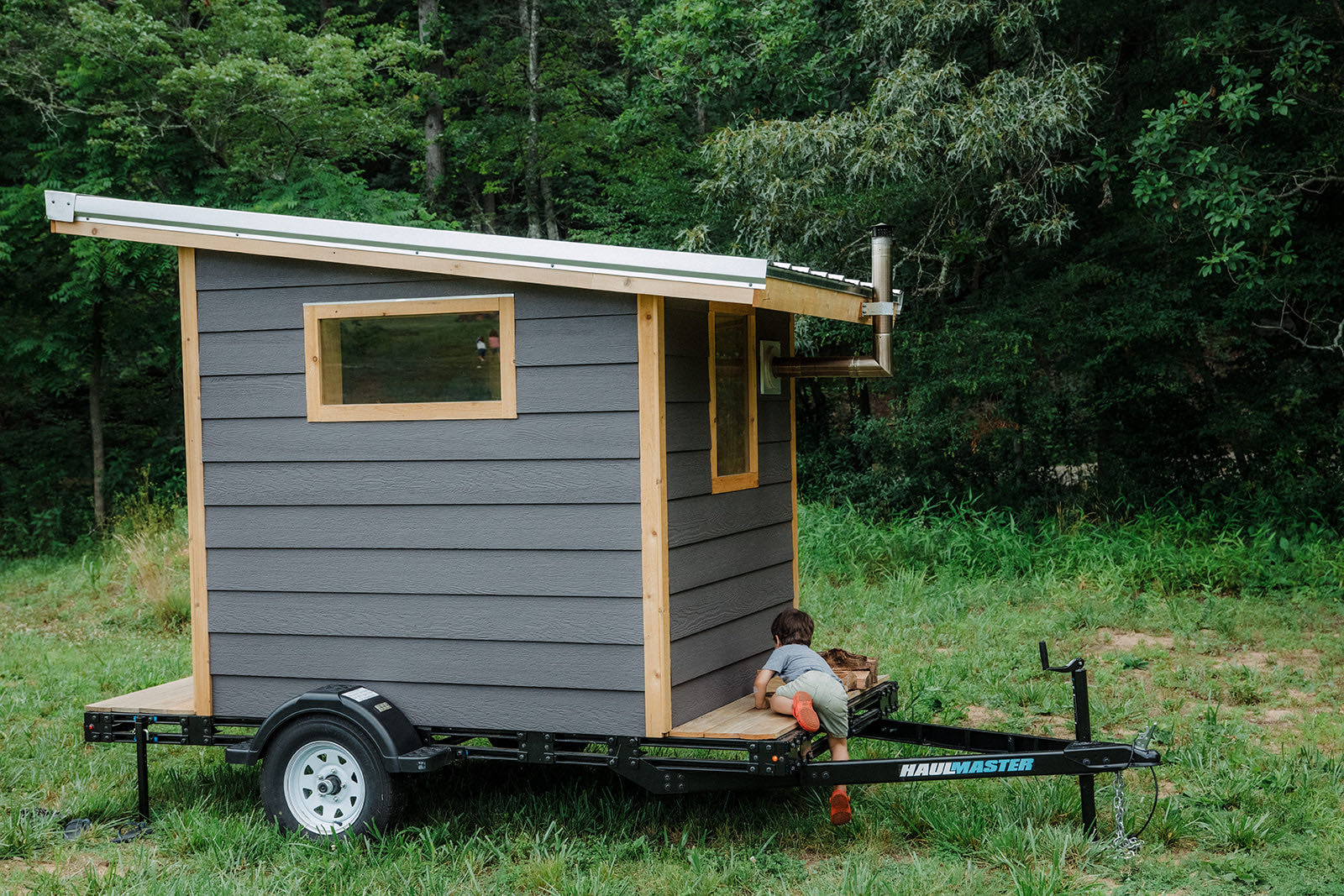 Full side profile of the Bushlot sauna on its utility trailer, charcoal lap siding with light wood corner trim and a single-slope roof