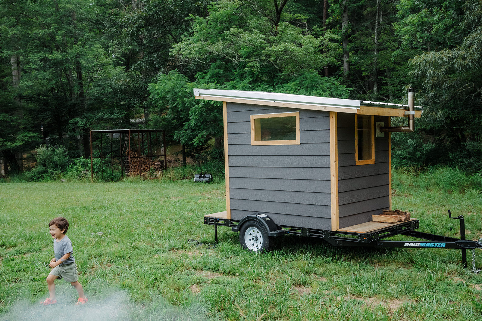 The finished sauna standing on its trailer in a mown field at the edge of the woods, steam drifting low across the grass as a small boy runs past