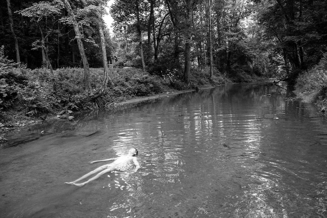 A swimmer floating on their back in a still forest creek, trees closing overhead