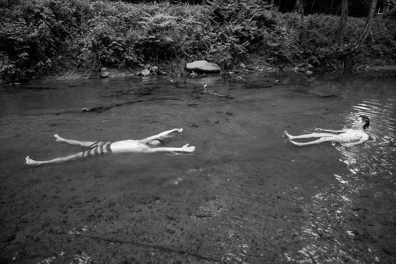 Two people floating on their backs in a shallow forest creek, arms out, looking up through the trees