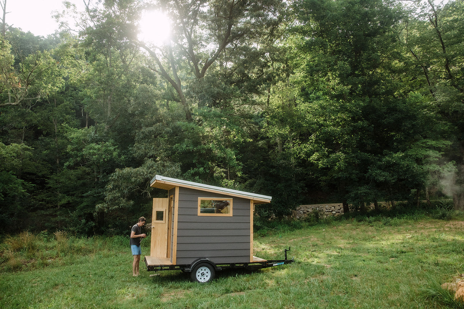 A Bushlot mobile sauna standing in a mown clearing at the edge of the woods, late sun breaking through the canopy, the builder at the open door