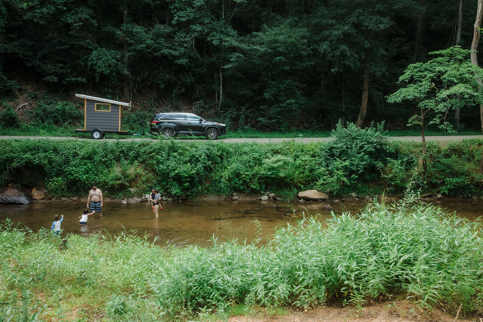 The sauna and its tow vehicle parked on a woodland road while a family wades in the creek below