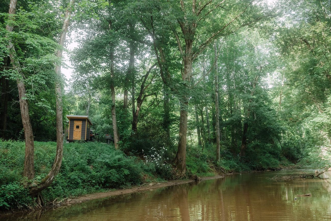 The sauna parked among tall hardwoods on the bank above a slow green creek