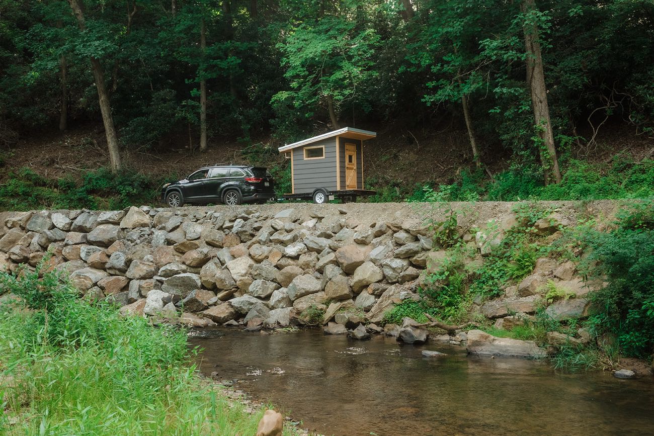 The Bushlot sauna towed behind an SUV along a gravel road above a dry-stacked stone wall and a shallow creek